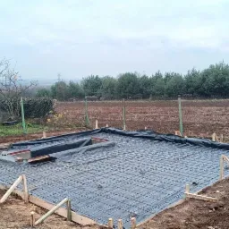 Przygotowanie zbrojenia pod płytę fundamentową, widoczne drewniane szalunki i folia izolacyjna, w tle dom i pole uprawne w pochmurny dzień.