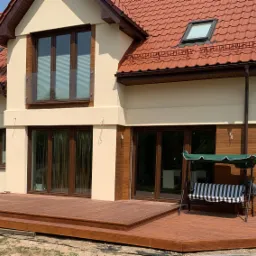 Nowoczesny dom z beżową elewacją, brązowymi oknami i dużym tarasem z drewna egzotycznego, na którym stoi huśtawka ogrodowa z zielonym daszkiem.