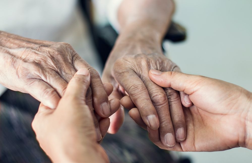 Dłoń osoby młodszej trzyma dłoń osoby starszej, widoczne zmarszczki na dłoni seniora, jasne tło.