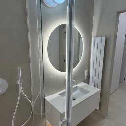 Kapibud - Nowoczesna łazienka z kabiną prysznicową ze szklanymi drzwiami, umywalką z szafką i okrągłym lustrem z podświetleniem LED. Minimalistyczny design, beżowe płytki.
