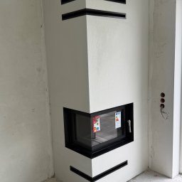 Kapibud - Nowoczesny narożny kominek z białej zabudowy z czarnymi poziomymi otworami wentylacyjnymi i przeszklonym wkładem, w pomieszczeniu w stanie deweloperskim.