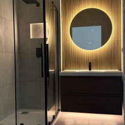 Kapibud - Elegancka łazienka z kabiną walk-in, ciemną szafką pod umywalkę i okrągłym lustrem z ciepłym podświetleniem na drewnianej ścianie lamelowej. Szare, wielkoformatowe płytki.