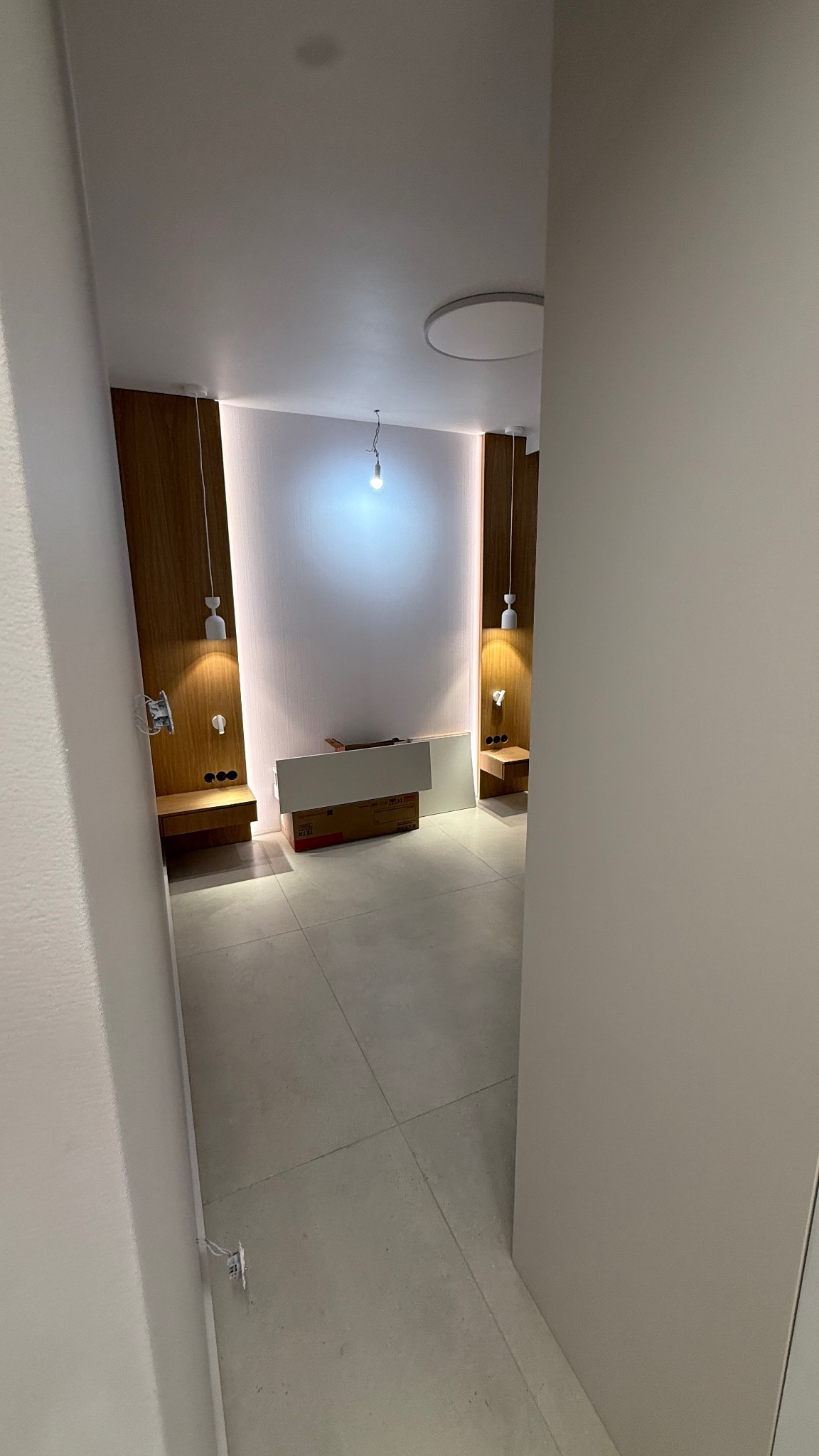 Wnętrze pokoju w trakcie wykańczania: szare płytki podłogowe, drewniane panele ścienne z oświetleniem, kartony i elementy wyposażenia. Widok zza framugi.