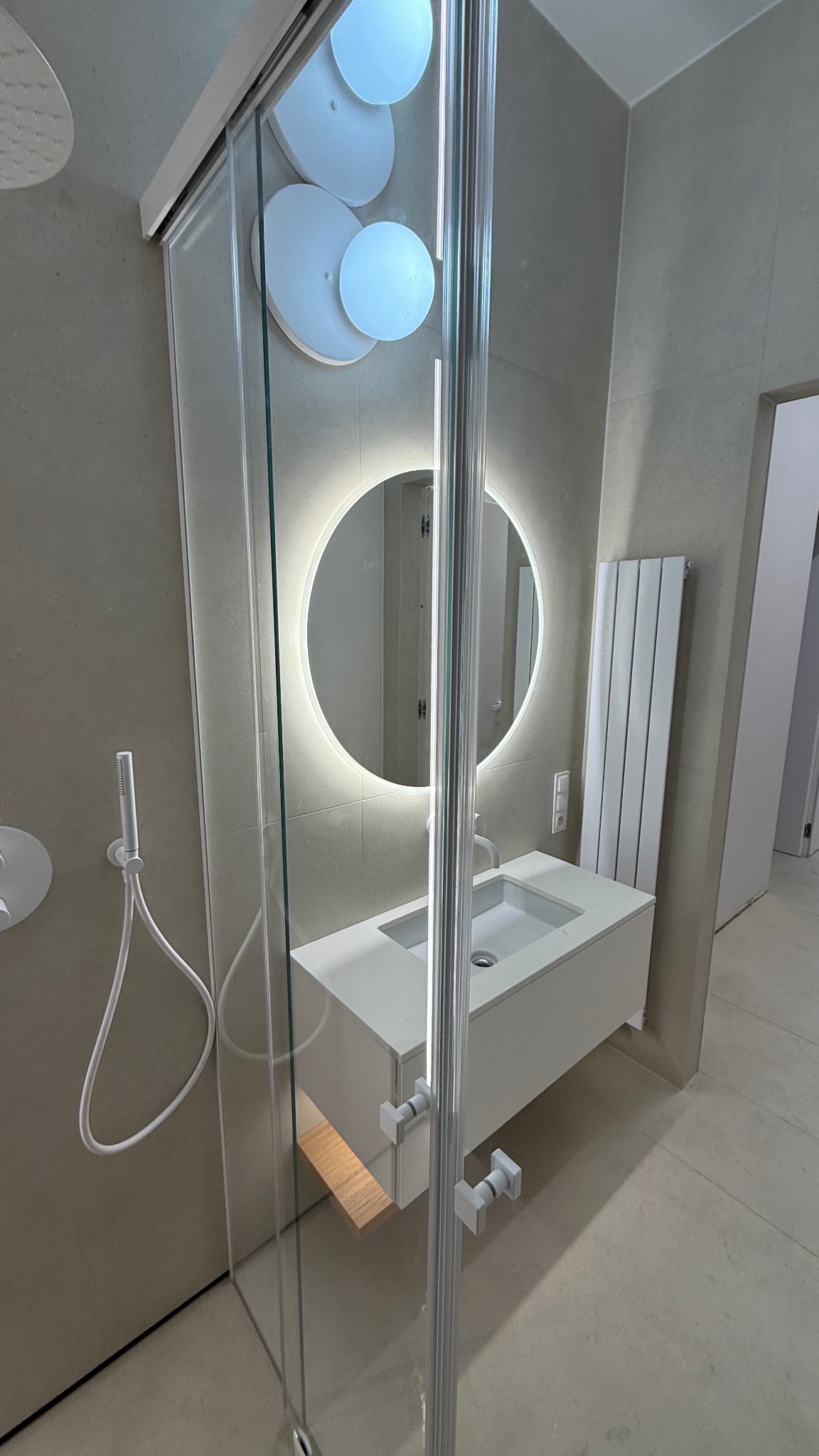 Nowoczesna łazienka z kabiną prysznicową ze szklanymi drzwiami, umywalką z szafką i okrągłym lustrem z podświetleniem LED. Minimalistyczny design, beżowe płytki.