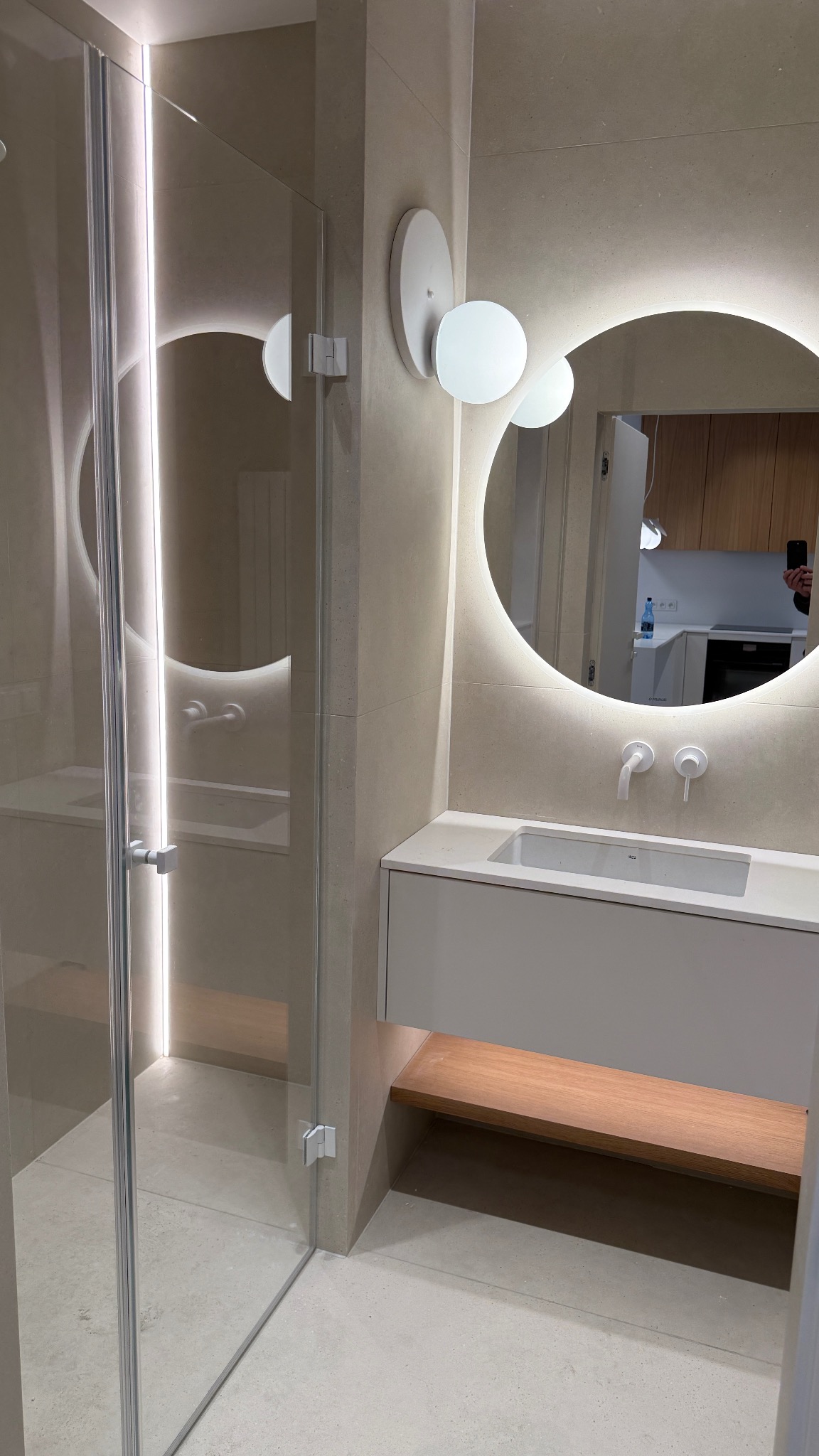 Nowoczesna łazienka z kabiną prysznicową ze szkła, umywalką z szafką i okrągłym lustrem z podświetleniem LED. Minimalistyczny design, beżowe płytki, drewniany akcent.