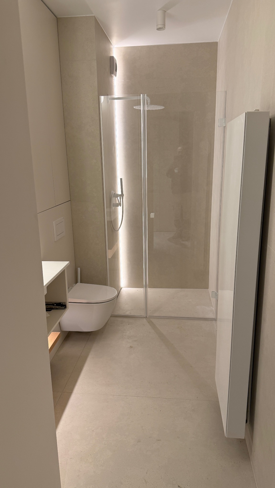 Nowoczesna łazienka z kabiną prysznicową typu walk-in, podwieszaną toaletą i grzejnikiem drabinkowym w minimalistycznym stylu, wykończona beżowymi płytkami.