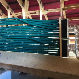 Montaż rekuperacji: Szereg zielonych, karbowanych rur wentylacyjnych ułożonych równolegle w przestrzeni poddasza, połączonych z metalową skrzynką i izolowanym kanałem wentylacyjnym. Widoczna...