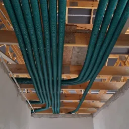 Zielone, karbowane rury wentylacyjne ułożone równolegle pod drewnianą konstrukcją sufitu w surowym wnętrzu budynku.