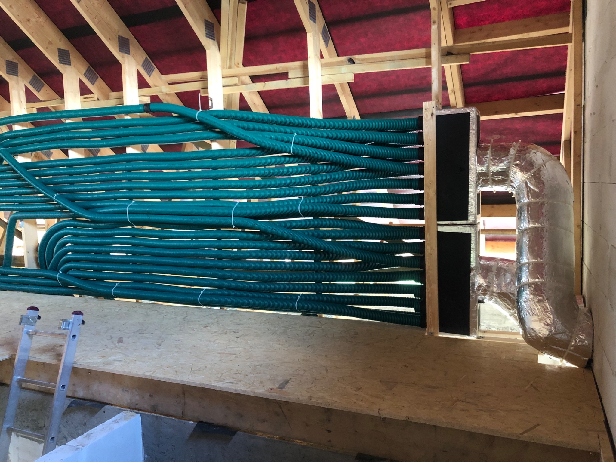 Montaż rekuperacji: Szereg zielonych, karbowanych rur wentylacyjnych ułożonych równolegle w przestrzeni poddasza, połączonych z metalową skrzynką i izolowanym kanałem wentylacyjnym. Widoczna...