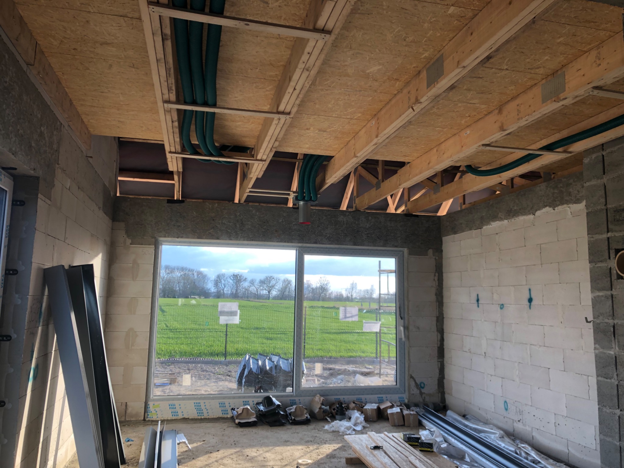 Wnętrze budynku w stanie surowym otwartym z widocznymi elementami instalacji wentylacyjnej w postaci zielonych rur podwieszonych pod stropem z płyt OSB. Widoczne okno z widokiem na zielone pole.