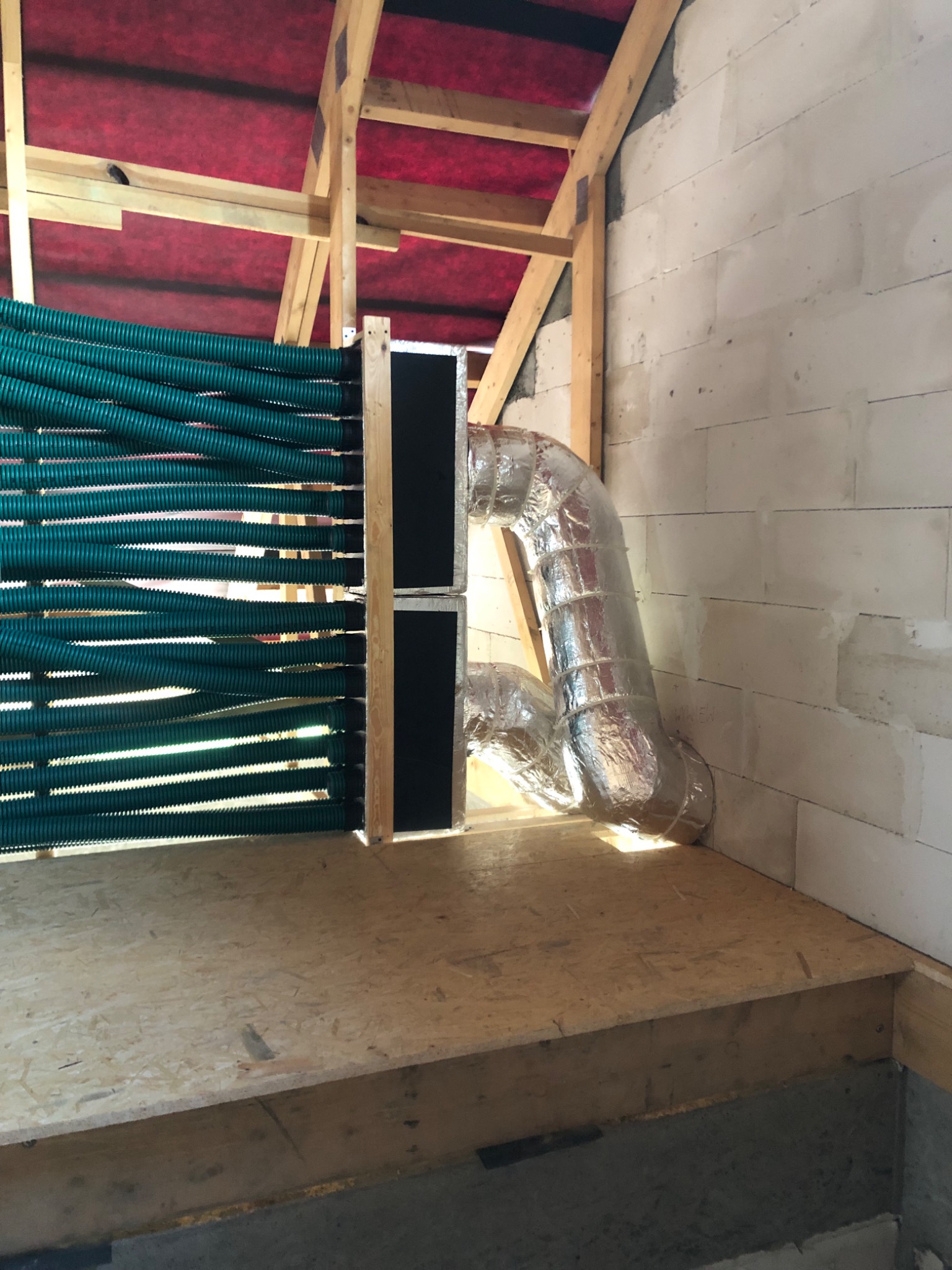 Wnętrze poddasza w trakcie prac instalacyjnych: widoczne zielone, karbowane rury wentylacyjne ułożone poziomo oraz srebrna, izolowana rura łącząca system, ściany z białych bloczków.