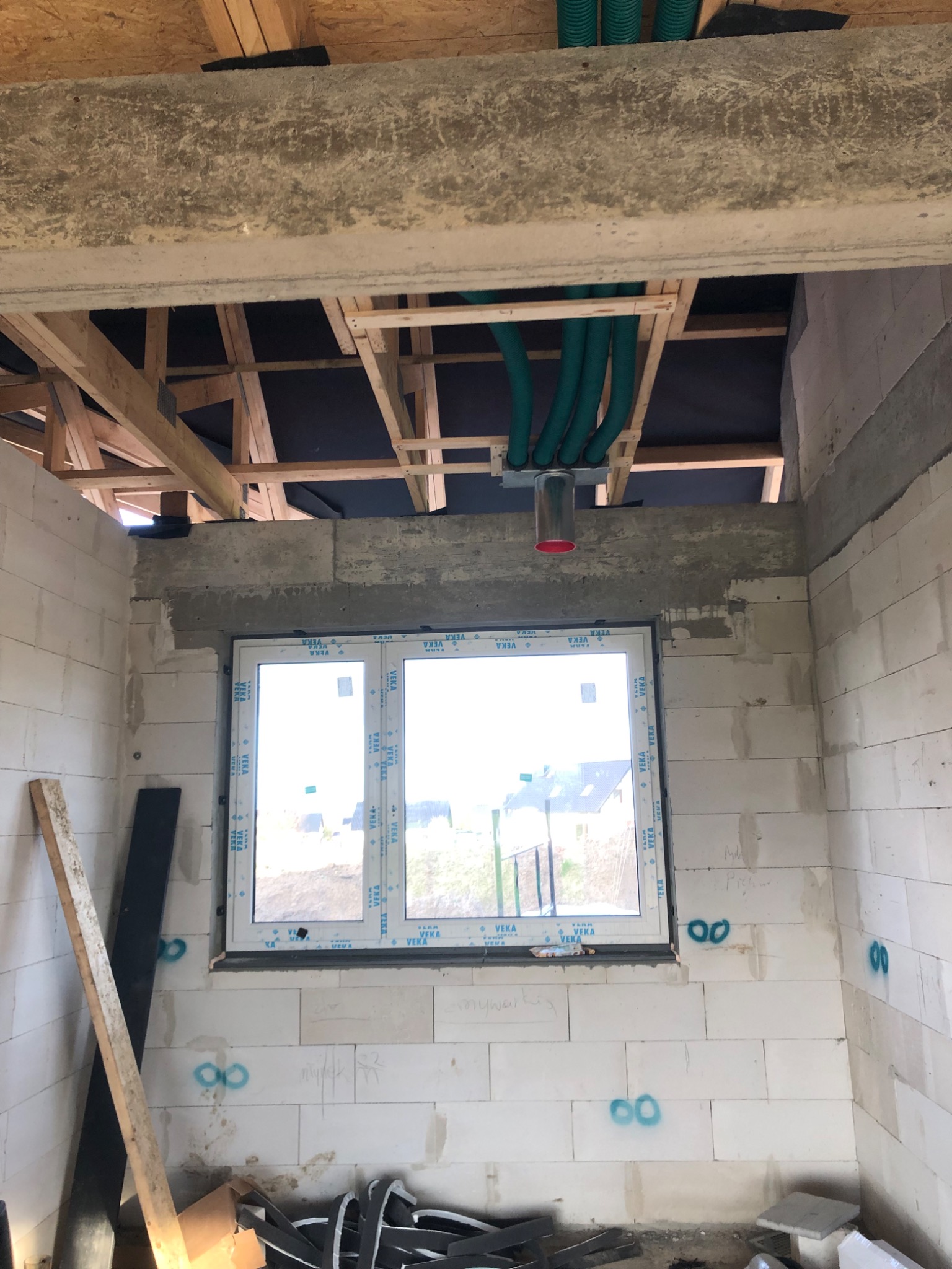 Surowy stan budynku w trakcie prac instalacyjnych: widoczne zielone rury wentylacyjne pod stropem, okno z PCV i ściany z białych bloczków z betonu komórkowego, na których widnieją oznaczenia...