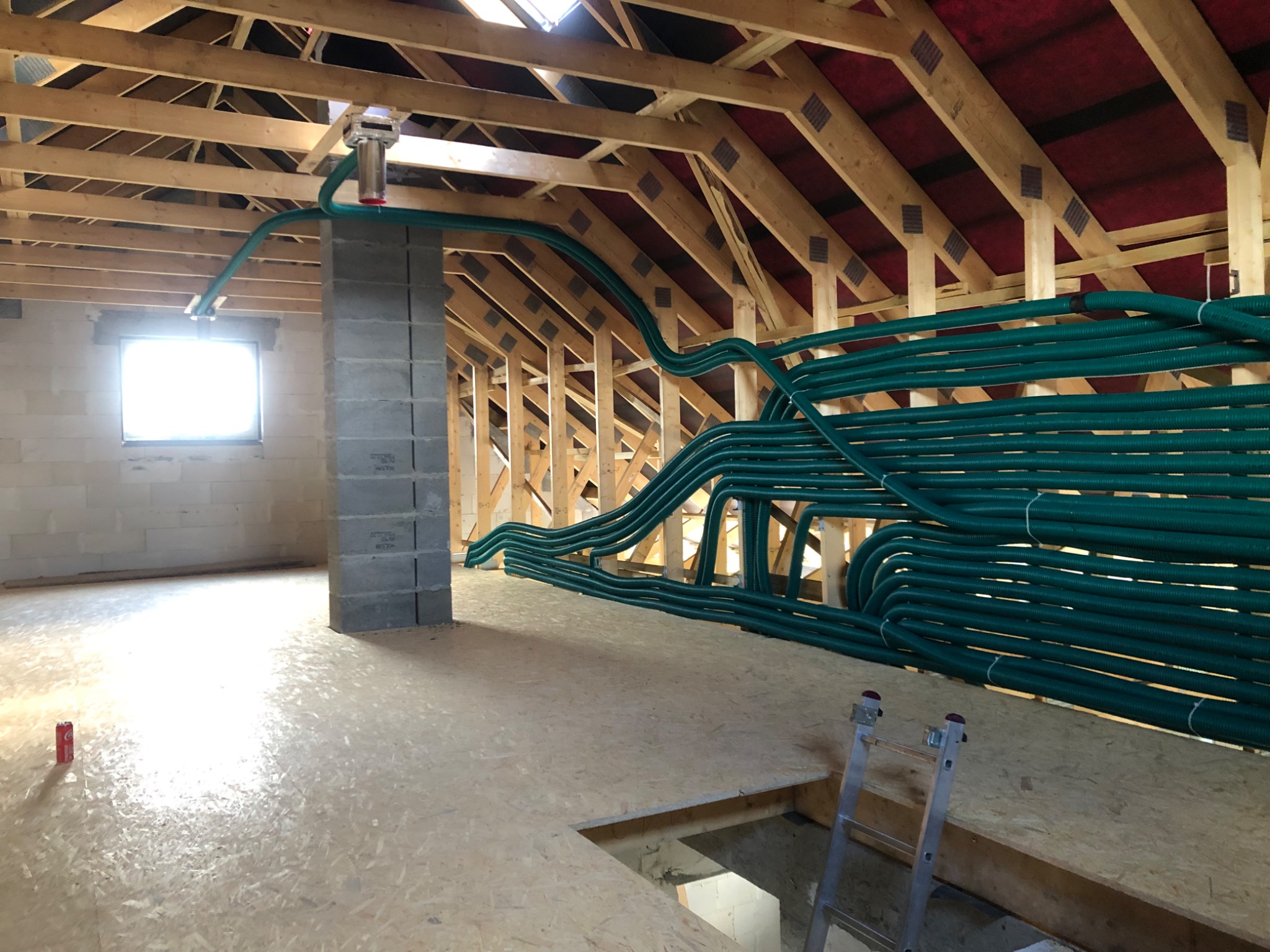 Zielone rury wentylacyjne ułożone na poddaszu w surowym stanie, widoczna konstrukcja dachu z drewnianych belek, okno i komin z pustaków, puszka po napoju na podłodze.