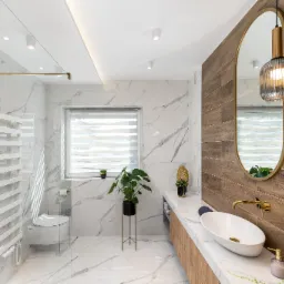 Nowoczesna łazienka z marmurowymi płytkami, drewnianymi akcentami, owalnym lustrem z wiszącymi lampami, białą umywalką nablatową i złotą armaturą.