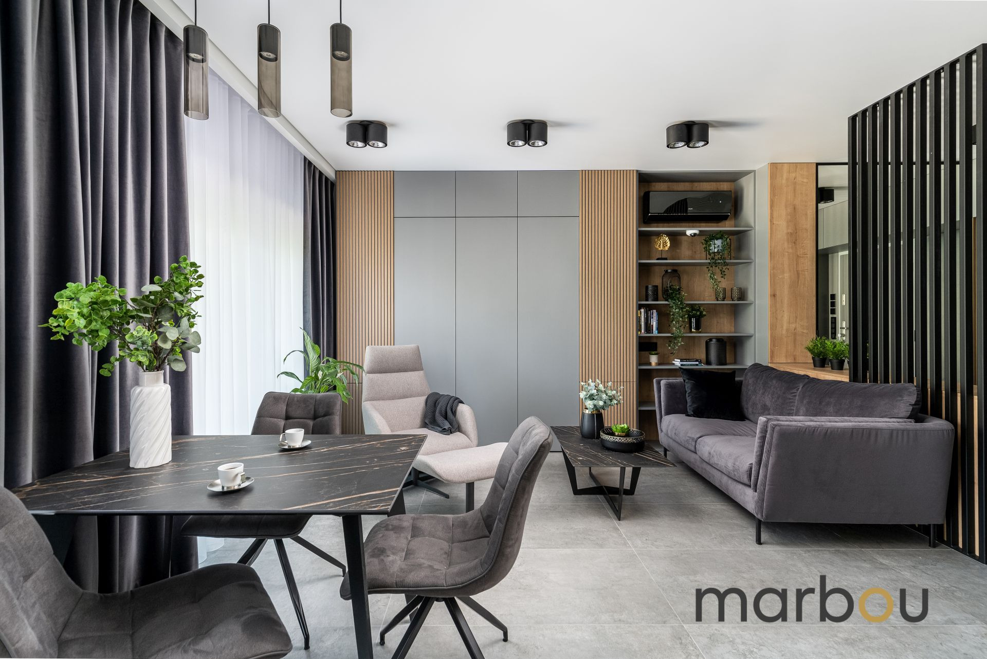 Nowoczesna aranżacja wnętrza mikroapartamentu: szary salon z marmurowym stołem, welurowe krzesła, minimalistyczne oświetlenie, drewniane panele ścienne i rośliny doniczkowe.