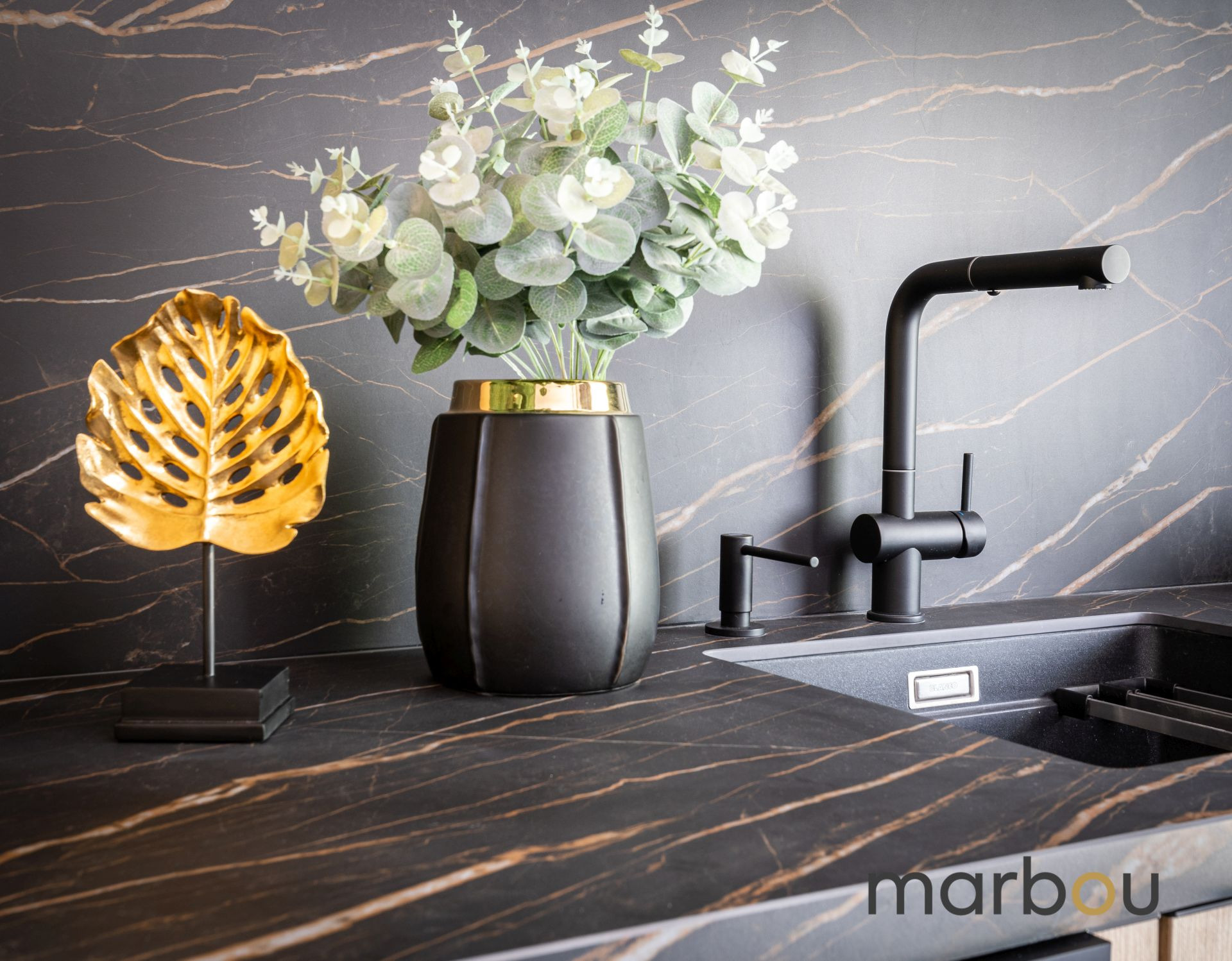 Czarna bateria kuchenna i zlew na blacie z ciemnego marmuru z dekoracjami: wazon z eukaliptusem i złoty liść monstery, tło stanowi ściana z marmuru.