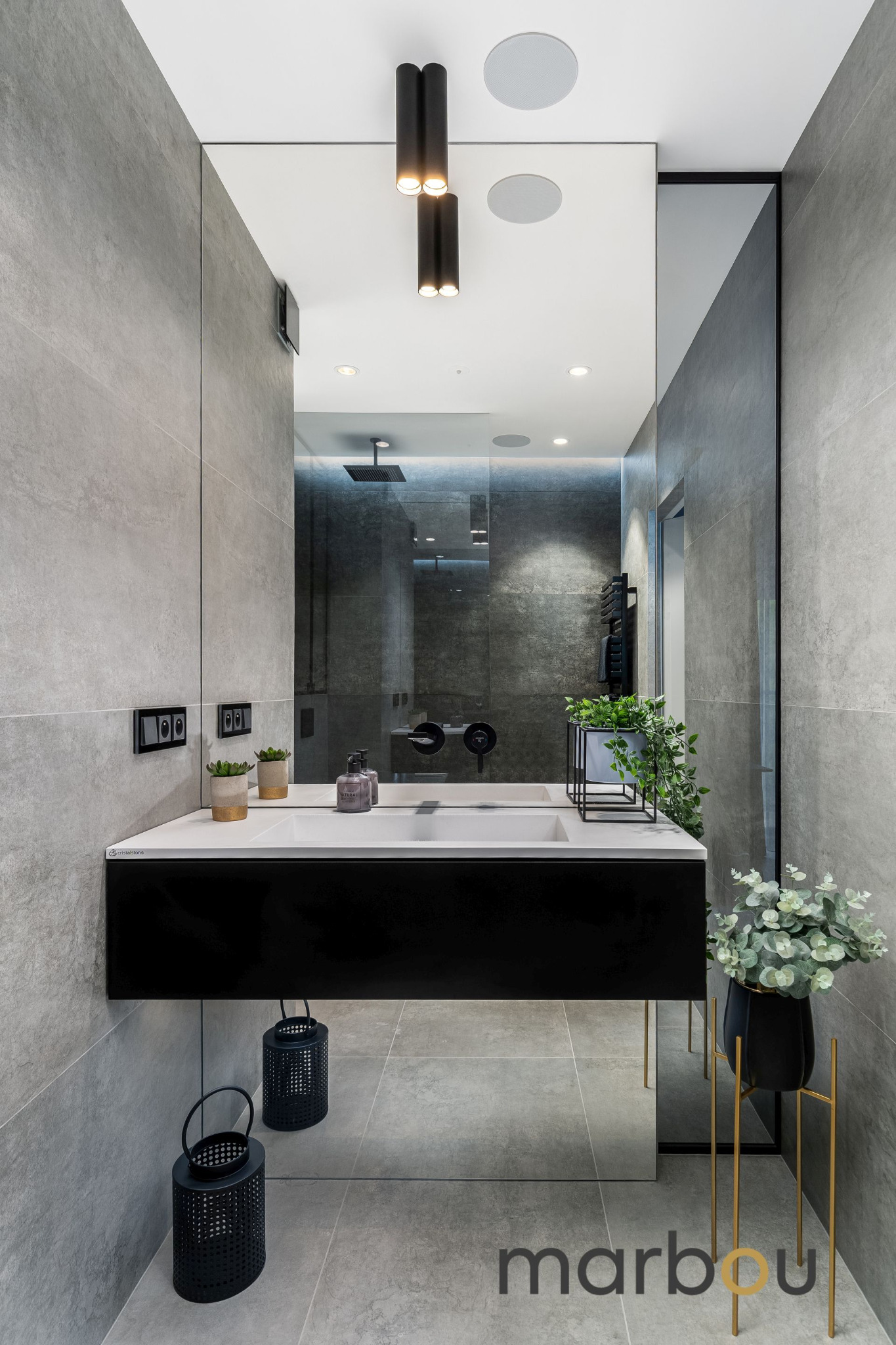 Nowoczesna łazienka w odcieniach szarości z czarnym blatem umywalki, minimalistycznymi lampami i roślinami doniczkowymi, widok na kabinę prysznicową.