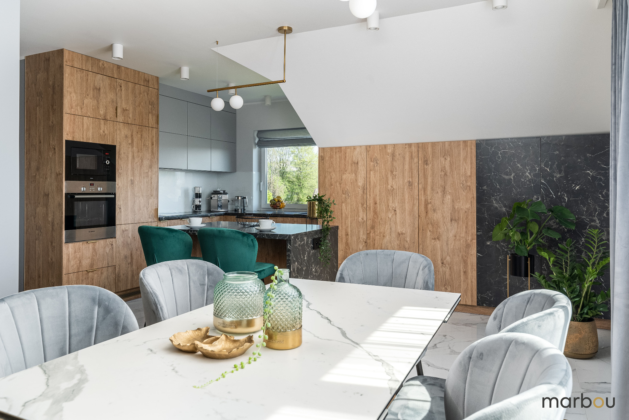 Jasne, nowoczesne wnętrze domu w Trzebini: marmurowy stół jadalny z dekoracjami, szare krzesła welurowe, drewniana zabudowa kuchenna z piekarnikiem i mikrofalówką, zielone krzesła barowe, okno...