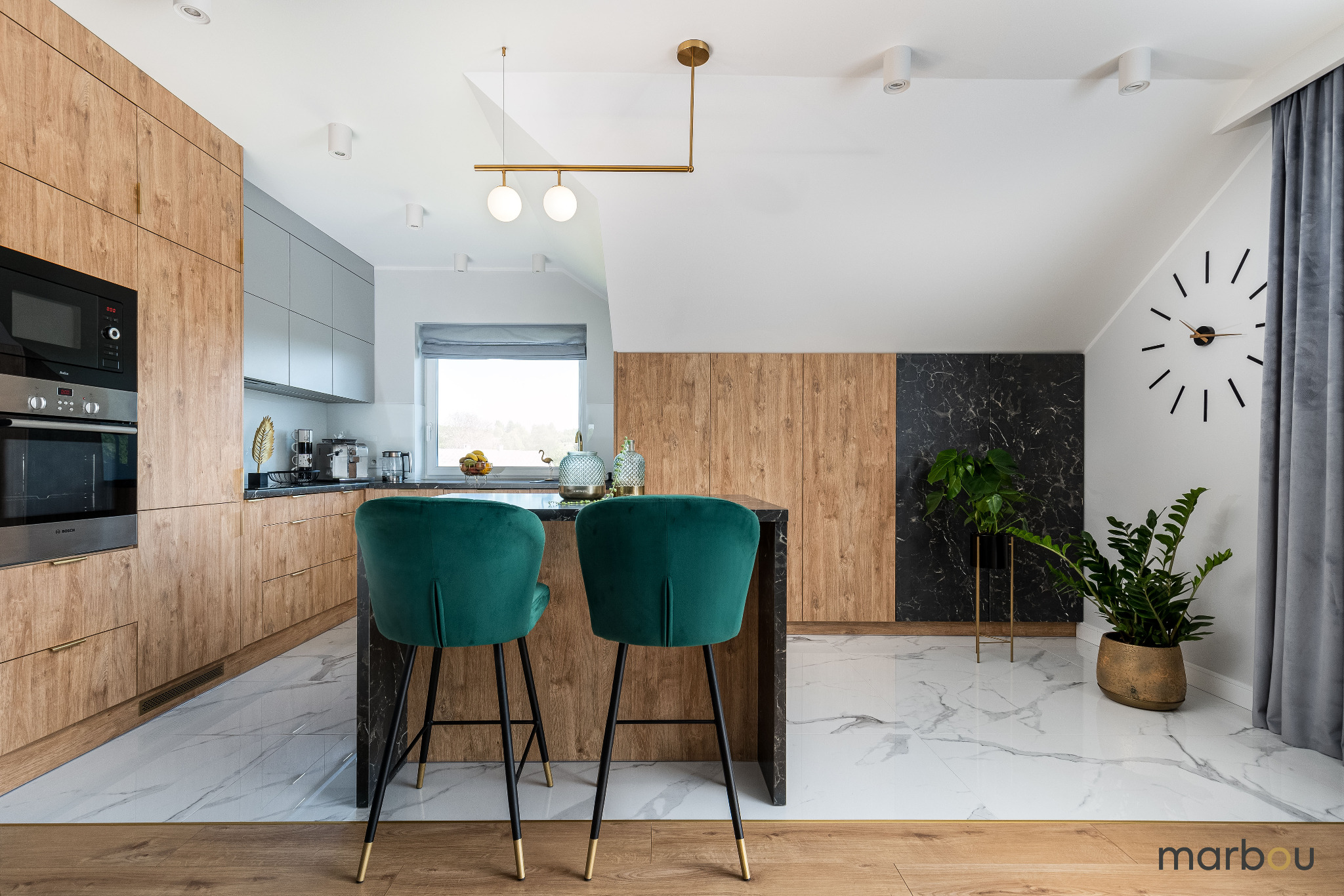 Nowoczesna kuchnia w domu w Trzebini: drewniane szafki, marmurowe podłogi, zielone hokery przy czarnym blacie wyspy kuchennej, złote detale oświetlenia i rośliny doniczkowe.