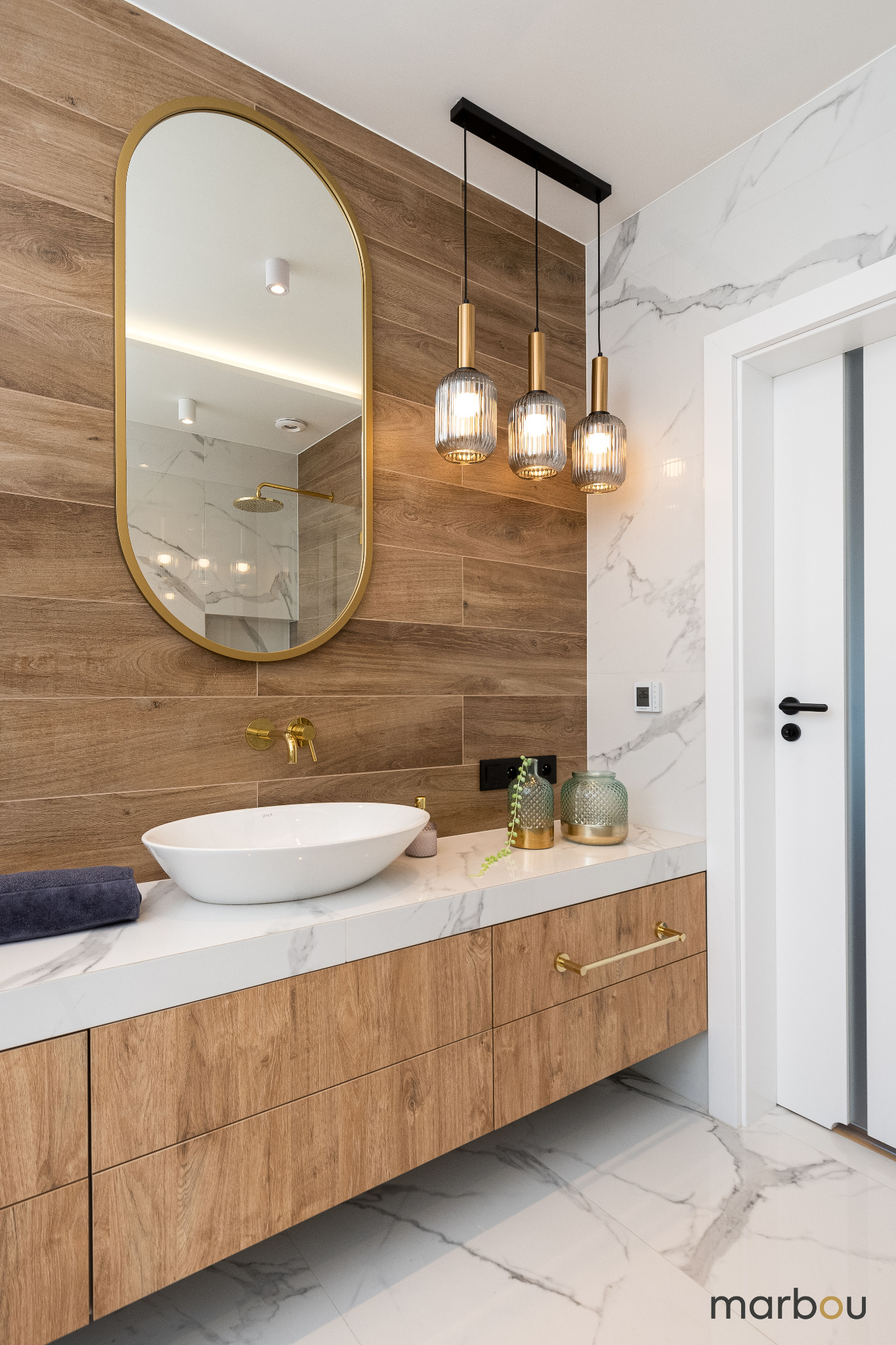 Nowoczesna łazienka z drewnopodobnymi płytkami, owalnym lustrem w złotej ramie, nablatową umywalką i marmurową podłogą. Wiszące lampy ze szklanymi kloszami.