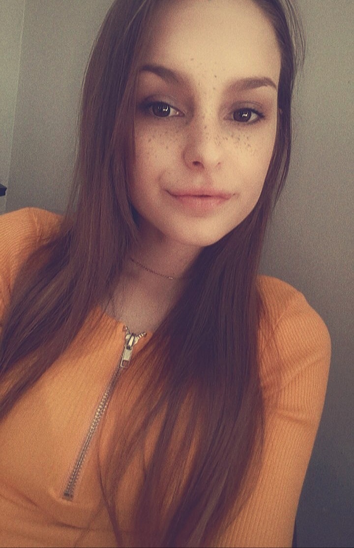 Portret młodej kobiety z piegami, uśmiechniętej, w pomarańczowej bluzce z suwakiem, na neutralnym tle.