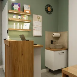 Recepcja w gabinecie kosmetycznym z drewnianym blatem, umywalką z kamienia i półkami z produktami kosmetycznymi na tle ściany w kolorze oliwkowym.