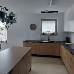 Jasna kuchnia z drewnianymi frontami szafek, czarnym blatem, białymi płytkami na ścianie i drewnianym zegarem. Na parapecie okna stoją doniczki z roślinami. Nad wyspą kuchenną wiszą rośliny...