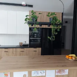 Nowoczesna kuchnia z drewnianymi szafkami, białymi płytkami na ścianie, czarnym sprzętem AGD i wyspą z owocami oraz dziecięcymi rysunkami.