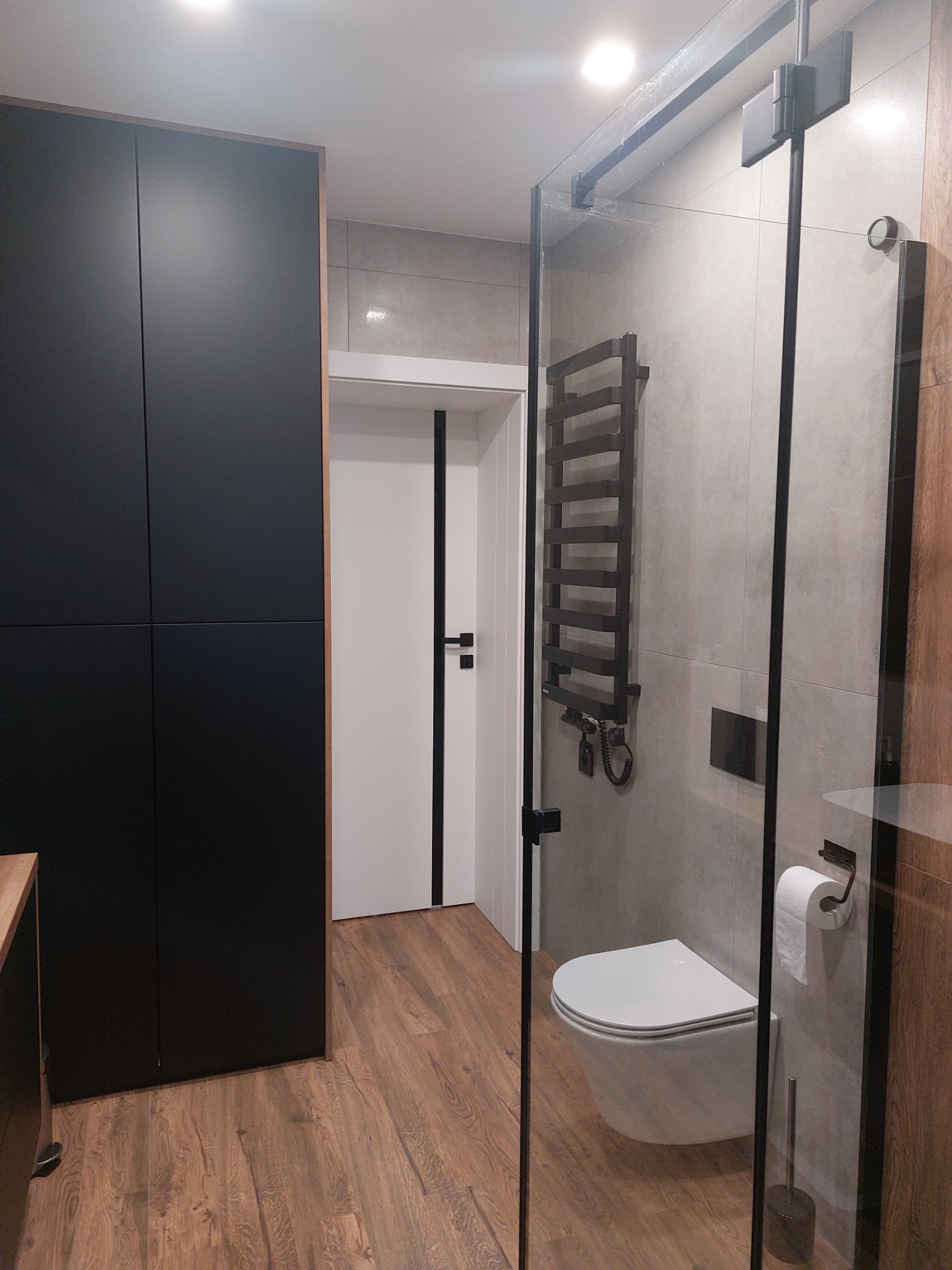 Nowoczesna łazienka z drewnianą podłogą, szarą glazurą, czarnym grzejnikiem drabinkowym i kabiną prysznicową ze szkła hartowanego, wisząca toaleta, minimalistyczna szafka i białe drzwi z czarnymi...
