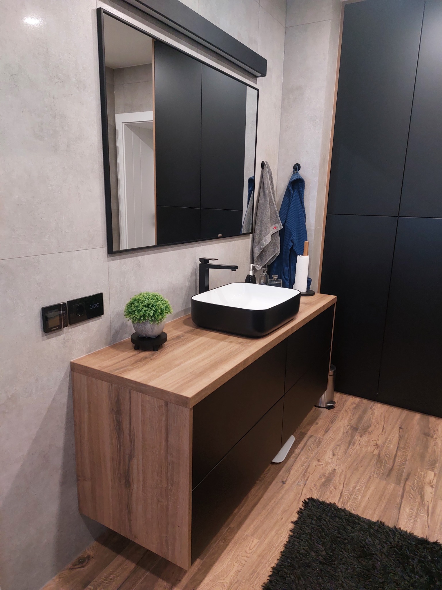 Nowoczesna łazienka z drewnianym blatem, czarną szafką pod umywalką, owalną umywalką nablatową, dużym lustrem w czarnej ramie, szarymi płytkami na ścianach, drewnopodobną podłogą i czarnym dywanikiem.