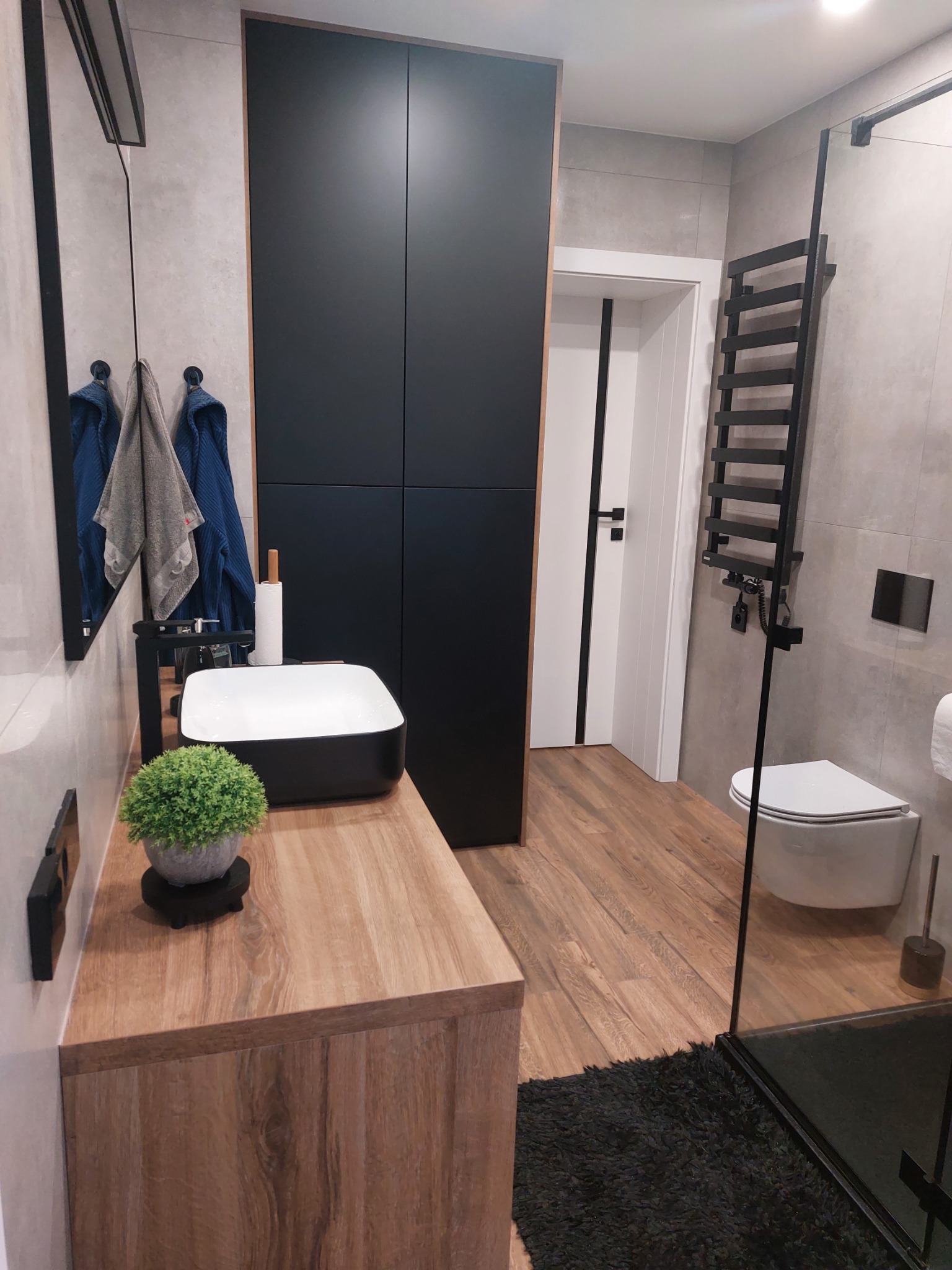 Nowoczesna łazienka w szarościach z drewnianym blatem, czarną szafką i umywalką nablatową, akcentem roślinnym w postaci małej doniczki oraz czarnym grzejnikiem drabinkowym.