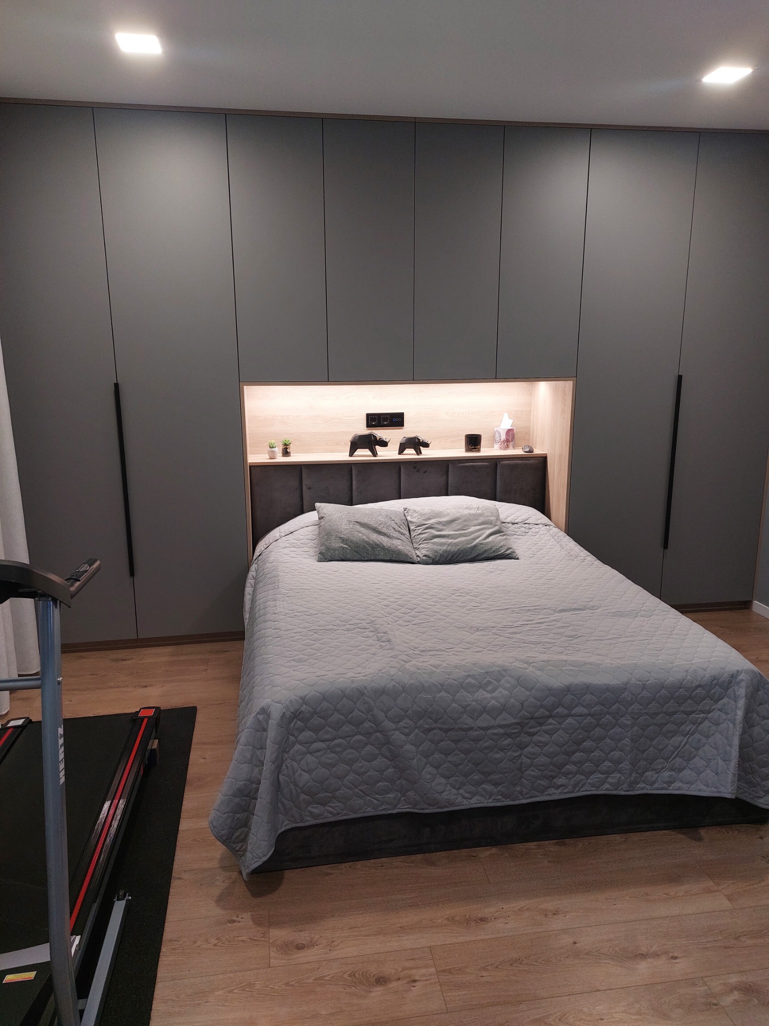 Szara sypialnia z łóżkiem z szarą narzutą, zabudową szaf i podświetlaną wnęką nad wezgłowiem, na półce dekoracje i roślinki, obok bieżnia.