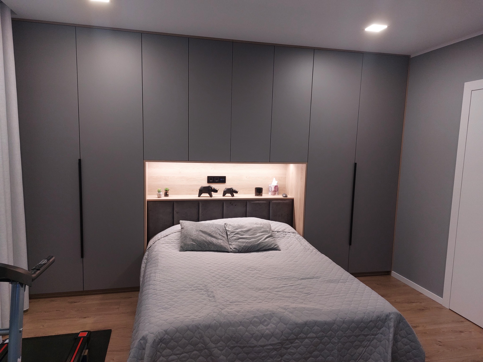Szara sypialnia z zabudową szaf na całej ścianie, oświetlona wnęka nad wezgłowiem łóżka z dekoracjami i roślinami, parkiet na podłodze, widoczny fragment bieżni.