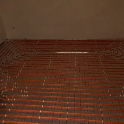 Pomarańczowe rurki ogrzewania podłogowego ułożone w pętle na siatce, przygotowane do zalania wylewką betonową w pomieszczeniu o surowych ścianach.