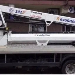 Podnośnik koszowy na samochodzie dostawczym, model 202R Revolution, z napisem 'Wynajmę' i numerem telefonu. Widok z boku na tle budynku z apteką.