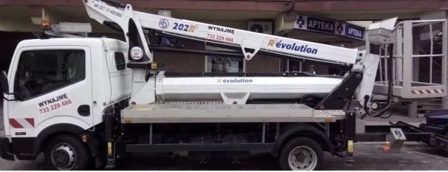 Podnośnik koszowy na samochodzie dostawczym, model 202R Revolution, z napisem 'Wynajmę' i numerem telefonu. Widok z boku na tle budynku z apteką.