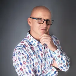 Portret łysiejącego mężczyzny w okularach i koszuli w roboty, z dłonią podpierającą brodę na szarym tle.