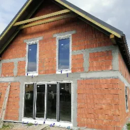 Surowy budynek z czerwonej cegły z zamontowanymi oknami, widoczne pianowanie i zabezpieczenia na ramach okiennych, drewniana konstrukcja dachu w trakcie budowy.