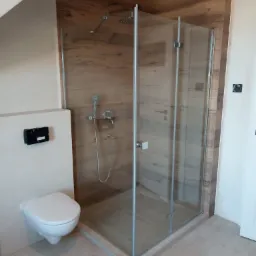 Nowoczesna kabina prysznicowa z deszczownicą, obudowana panelami imitującymi drewno, z wiszącą toaletą i dekoracyjną podłogą.