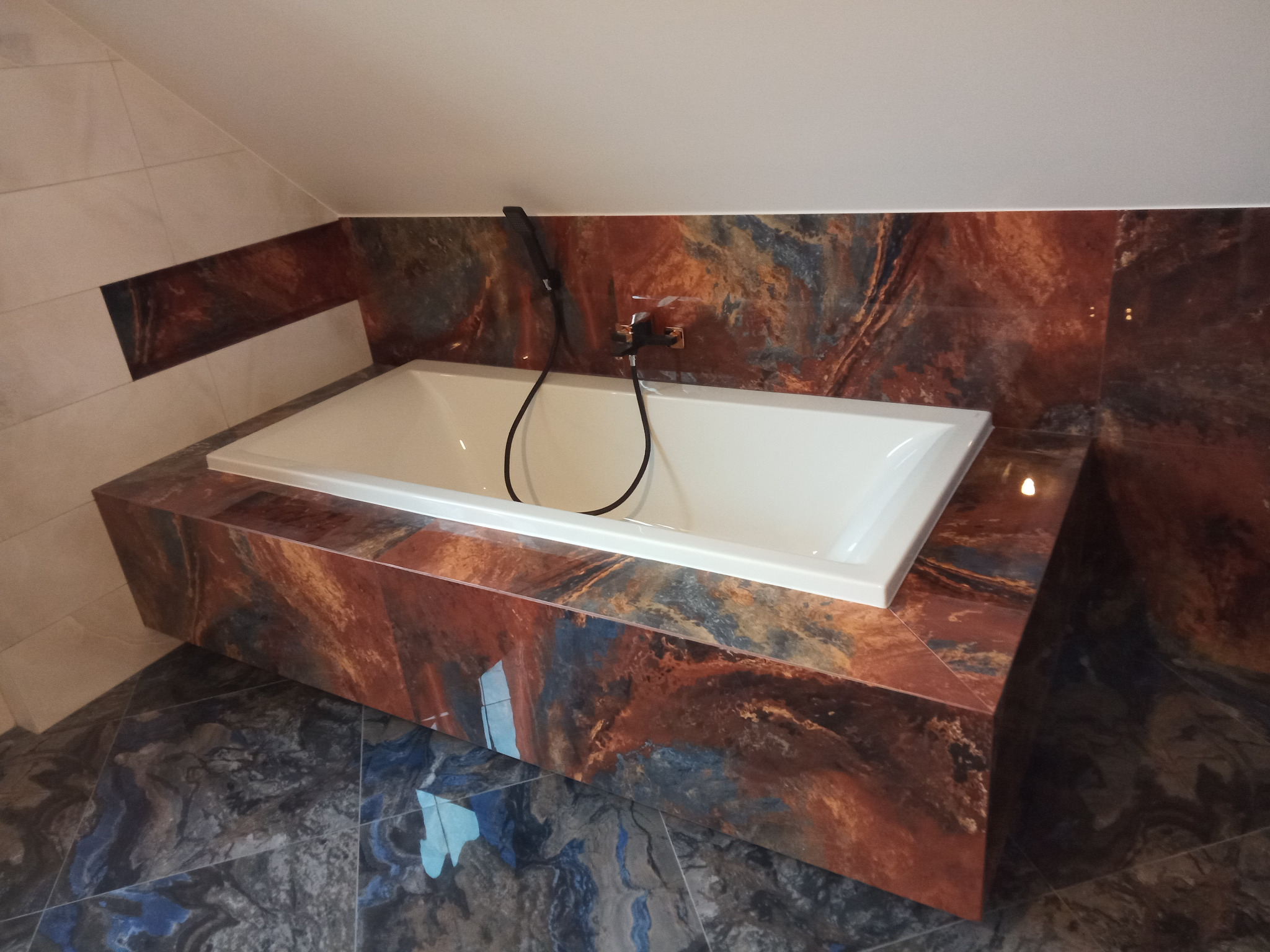 Biała wanna w obudowie z płytek imitujących marmur w odcieniach brązu i niebieskiego, dopasowane płytki podłogowe, bateria natryskowa w kolorze czarnym.