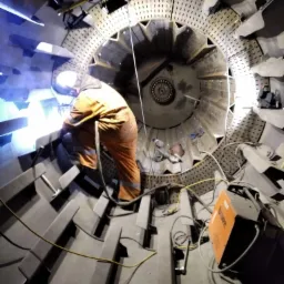 Spawacz w pomarańczowym kombinezonie spawa metalową konstrukcję wewnątrz dużej, okrągłej obudowy z licznymi otworami i elementami przypominającymi łopatki, widoczna spawarka obok.