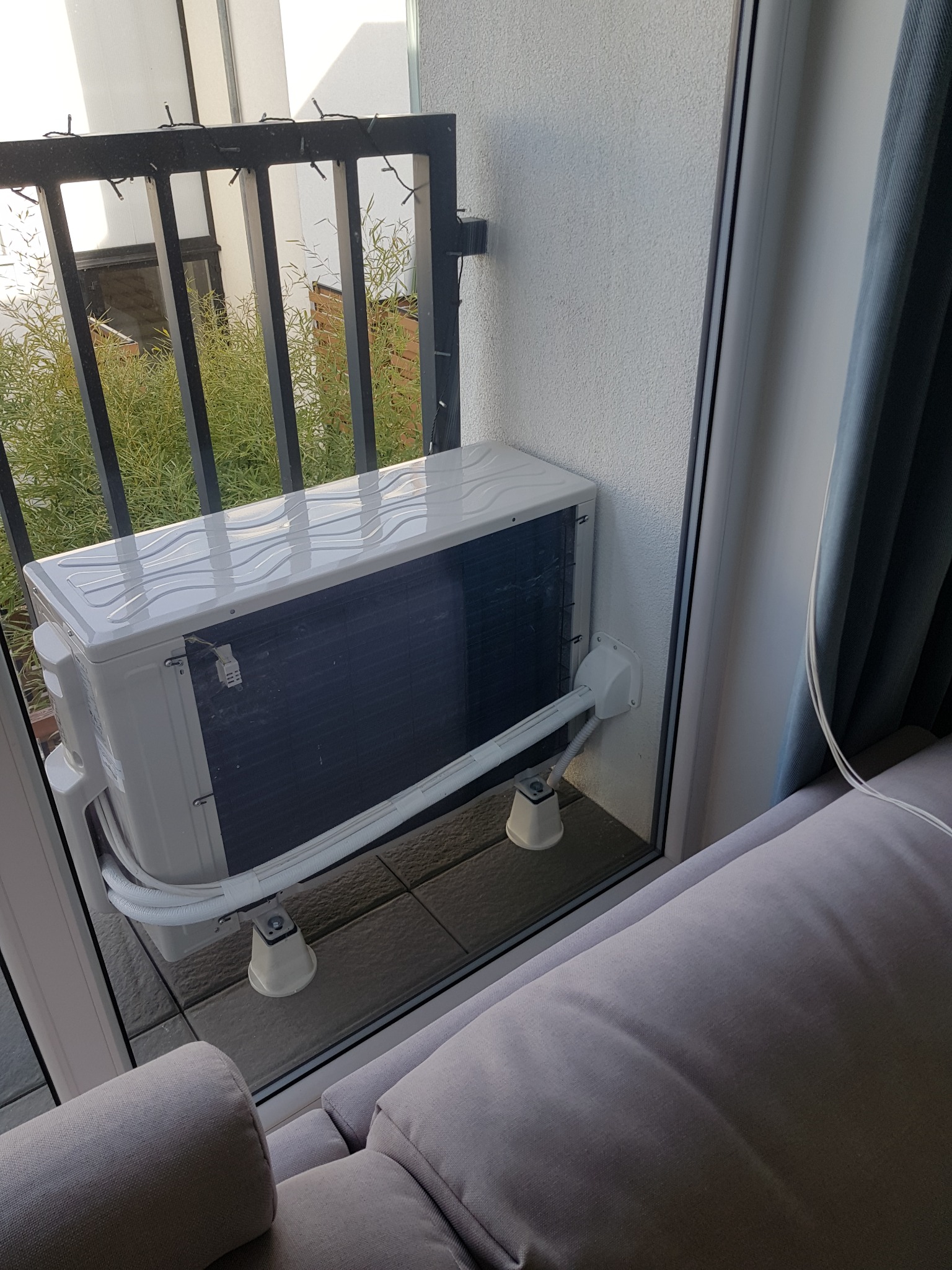 Zewnętrzna jednostka klimatyzacji zamontowana na balkonie, widoczna z wnętrza mieszkania, z fragmentem sofy w kadrze.