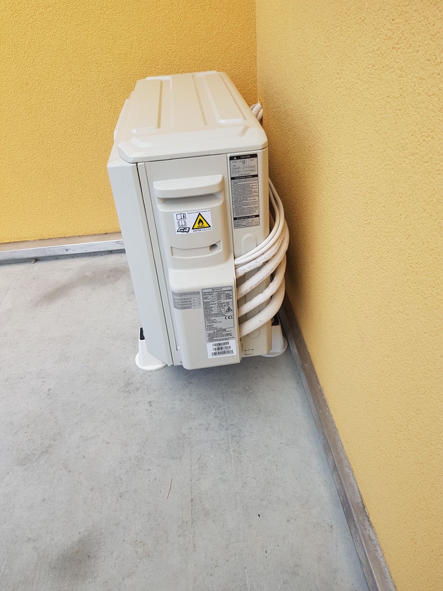 Zewnętrzna jednostka klimatyzacji zamontowana przy żółtej, teksturowanej ścianie budynku, widoczne przewody i naklejki ostrzegawcze.
