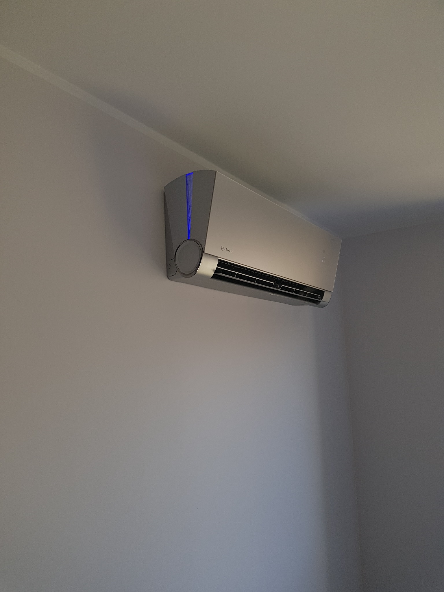 Nowoczesny klimatyzator ścienny w kolorze srebrnym z niebieskim akcentem, zamontowany w rogu pokoju na gładkiej, szarej ścianie.