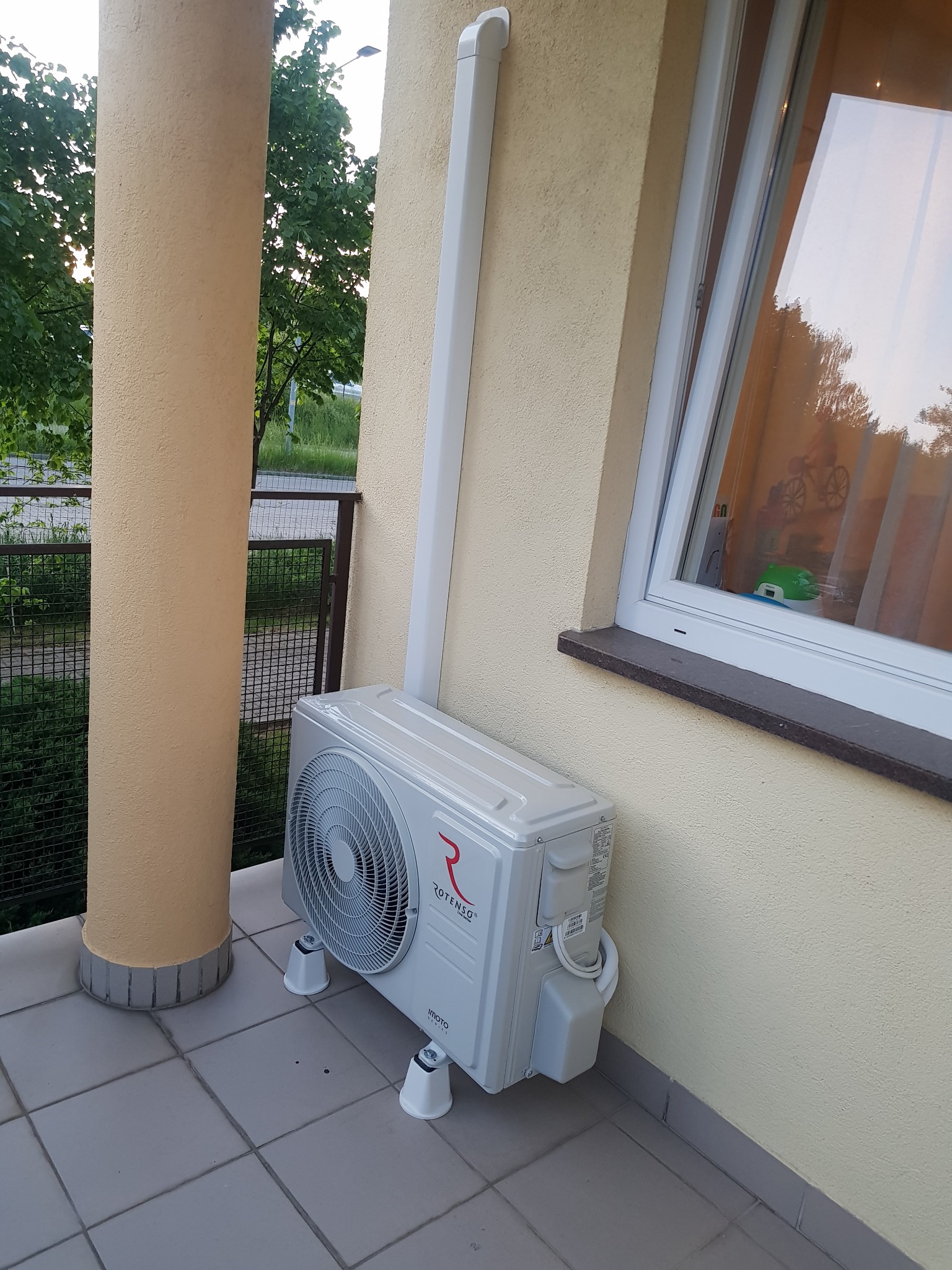 Zewnętrzna jednostka klimatyzacji Rotenso Imoto Plus zamontowana na balkonie, na białych nóżkach, z rurą odprowadzającą skropliny przy ścianie, widok na kolumnę balkonu i okno z zabawkami w tle.