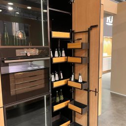 Nowoczesna zabudowa kuchenna z piekarnikiem i systemem cargo z półkami na butelki. Fronty w odcieniach drewna i czerni. Oświetlenie LED wewnątrz szafek.