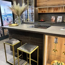 Nowoczesna kuchnia z wyspą, marmurowym blatem i złotymi akcentami. Hoker z szarym siedziskiem i złotą ramą. Podświetlane witryny eksponują dekoracje.