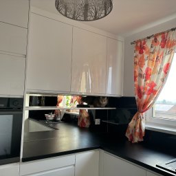 Biała, nowoczesna kuchnia z czarnym blatem i błyszczącymi szafkami. W lustrzanej przestrzeni między szafkami siedzi kot. Okno z zasłonką w kwiaty i wazonem z tulipanem.