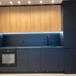 Nowoczesna kuchnia z ciemnymi szafkami i drewnianymi górnymi frontami, oświetleniem LED pod szafkami, z piekarnikiem i zlewozmywakiem. W tle wzorzysta tapeta.