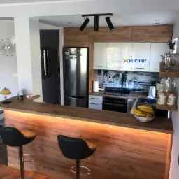 Nowoczesna kuchnia z drewnianym blatem barowym podświetlonym od spodu, dwa czarne hokery na chromowanych nogach, na ścianie fototapeta z motywem miasta.